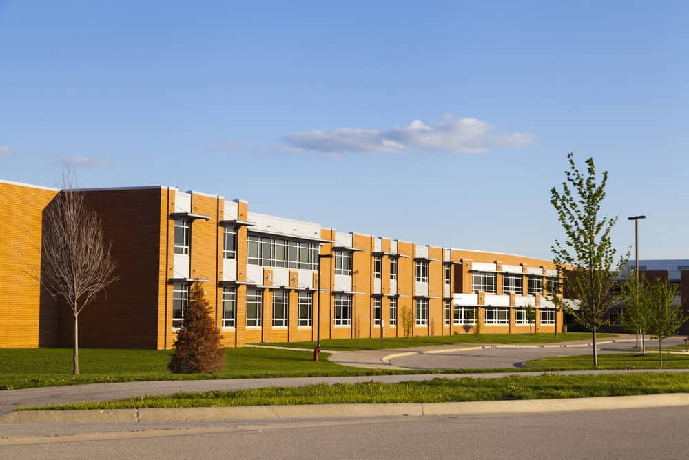 The exterior of the two-story brick school building, boasting large windows and surrounded by lush grass and a few young trees, shines under the clear blue sky. Attentive school lawn care ensures a pristine environment for students and faculty alike.