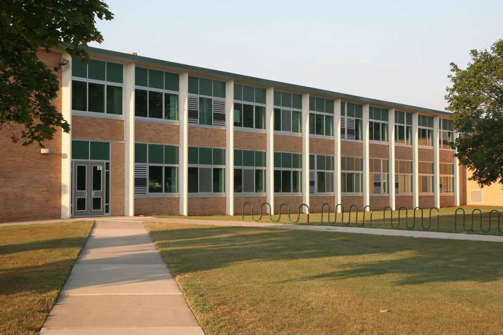 A three-story brick building with green and white window frames serves as a charming university hub. A well-manicured lawn cared for by professionals lines the sidewalk leading to the entrance, mingled with trees that offer a welcoming shade.