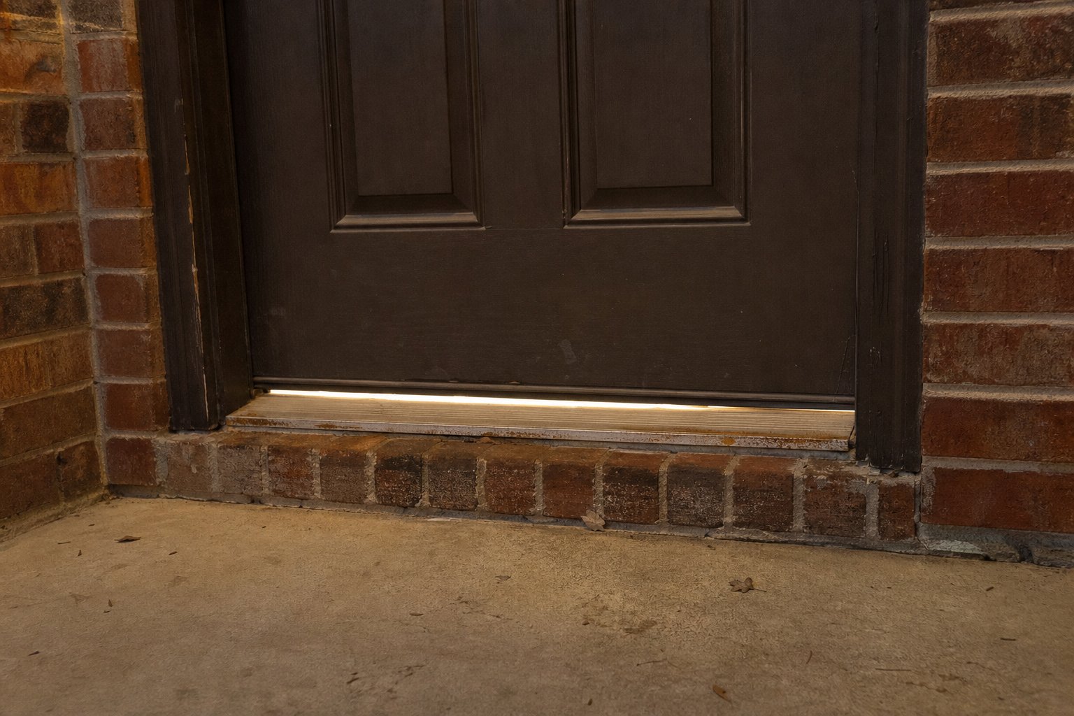 Front door threshold of a brick home with worn weatherstripping and a small gap of light showing underneath, evening porch light glow