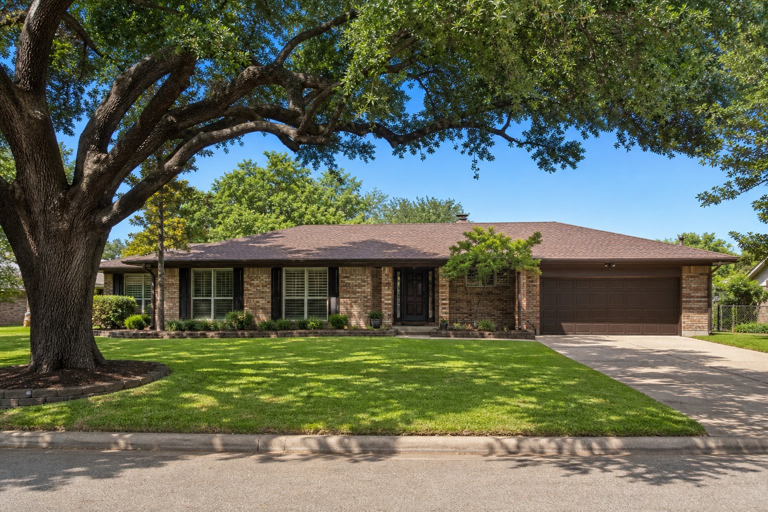 Well-kept single-story brick ranch home on a slab foundation with healthy green lawn and mature live oak, clear summer sky, North Texas suburban street