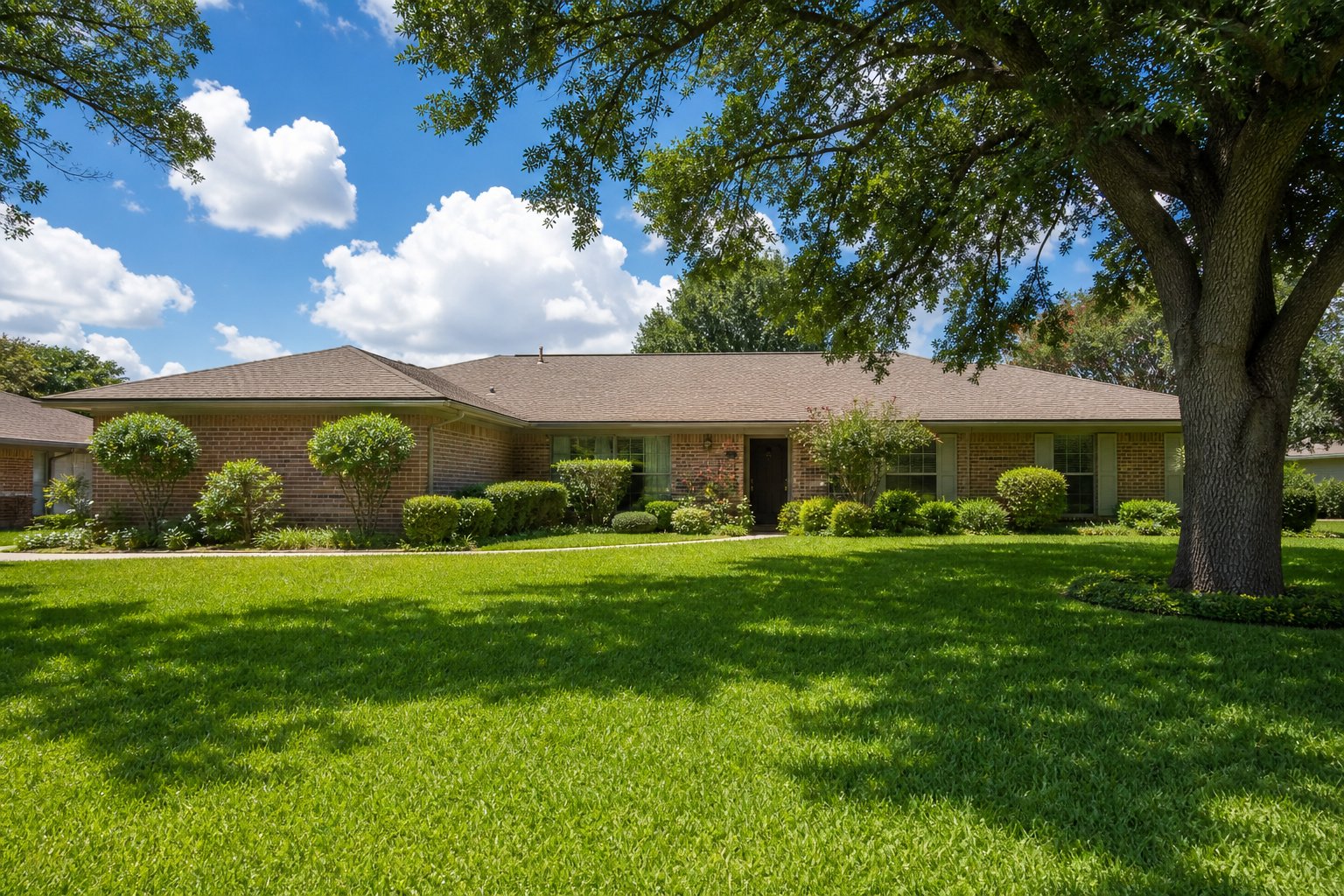 Tidy single-story brick ranch home with lush green St. Augustine lawn under a big summer sky, mature shade oaks, North Texas suburban neighborhood