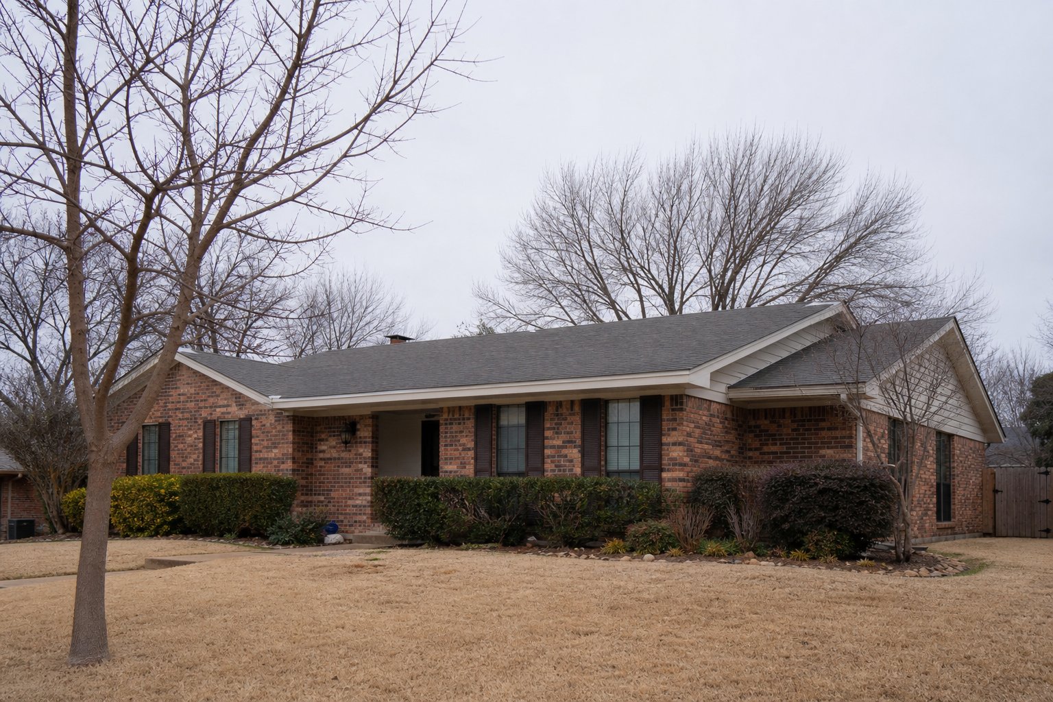 Brick ranch home in winter, dormant tan Bermuda lawn and bare tree branches against a pale gray sky, quiet late afternoon winter light