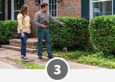 Two people stand outside a brick house, engaged in conversation about the neatly trimmed bushes along the walkway, while considering home pest control options to maintain their garden's health.