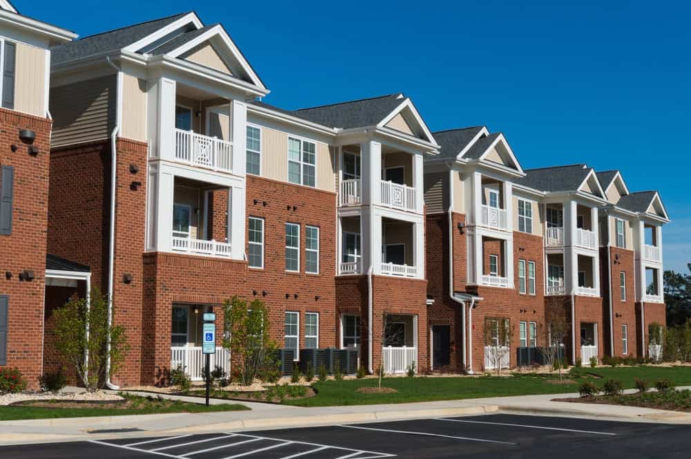 Three-story brick apartment building with white trim, balconies, and meticulously designed apartment landscaping. Parking lot with a designated accessible space in the foreground under a clear blue sky.