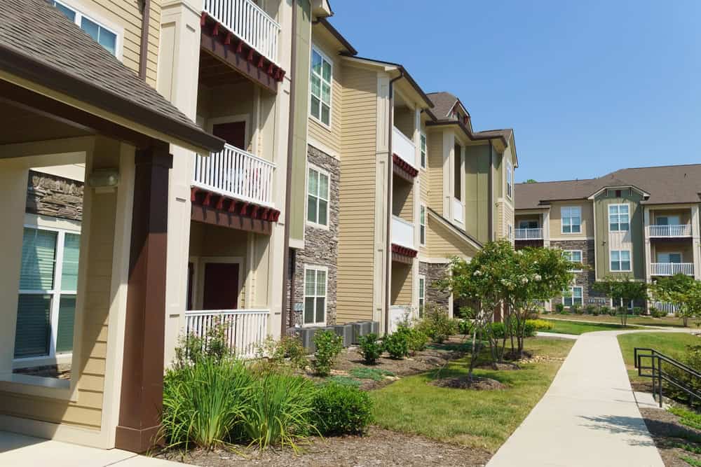 A complex of modern beige apartment buildings with balconies, surrounded by expertly designed apartment landscaping and a concrete walkway, under a clear blue sky.