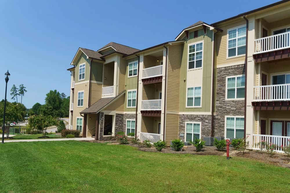 Three-story apartment building with beige siding, stone accents, and balconies features thoughtfully designed landscaping. Surrounded by a grassy lawn and sidewalk under a clear blue sky, this residence offers serene living.