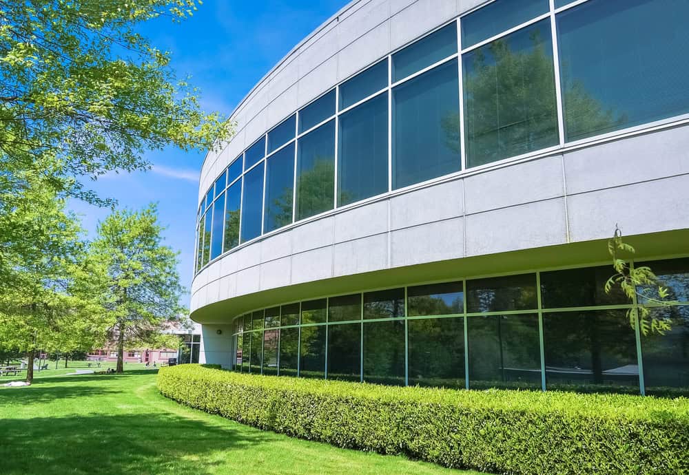 A modern building with large windows and a curved facade is surrounded by expertly maintained greenery, showcasing the benefits of commercial landscaping services under a clear blue sky.