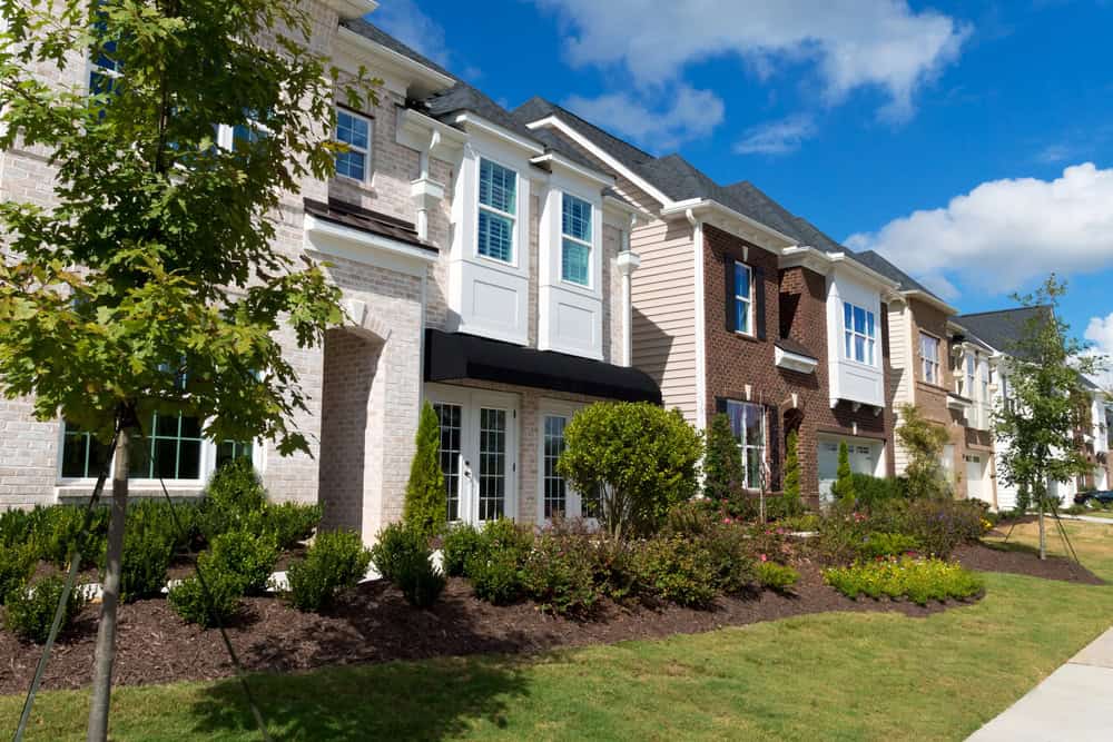 A row of modern townhouses with brick and siding exteriors, enhanced by meticulous apartment landscaping, is surrounded by lush gardens and a clear blue sky.