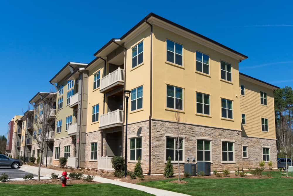 A modern three-story apartment building with yellow siding and stone accents, enhanced by meticulous landscaping. Its lush lawns and trees create a serene environment under a clear blue sky.