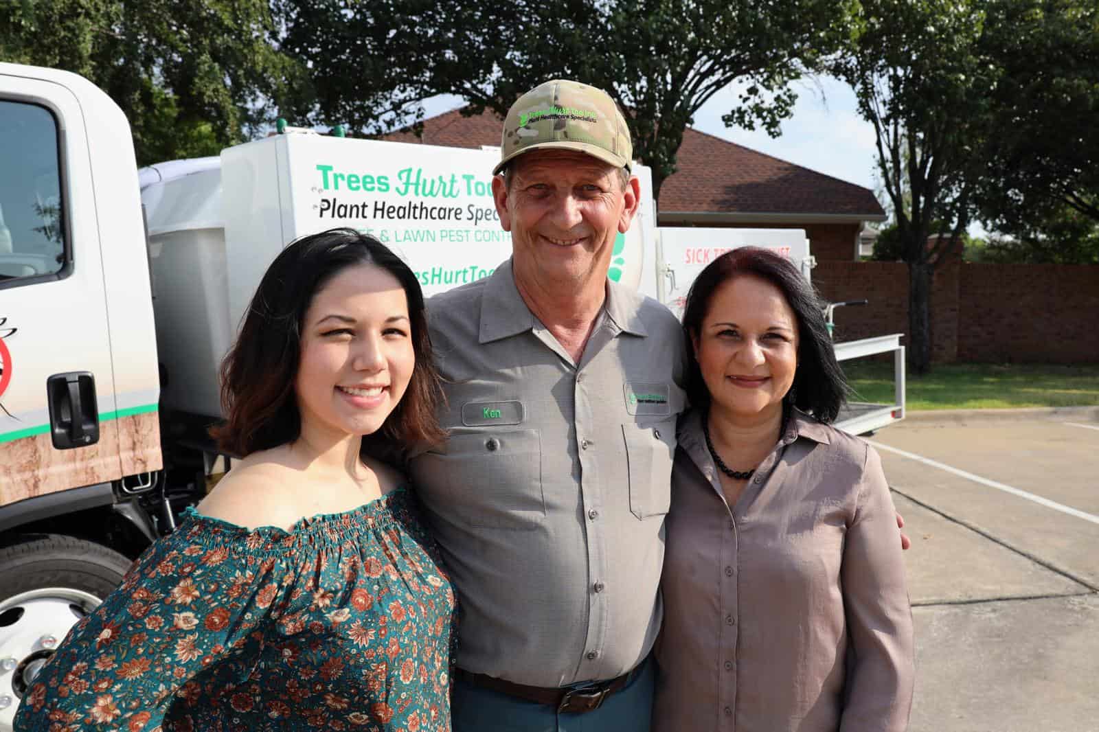 Three people smiling in front of a Trees Hurt Too, Inc. service truck proudly display their dedication as Plant Healthcare Specialists. With a backdrop of lush trees and a building, this scene perfectly encapsulates the essence of our About Us story.