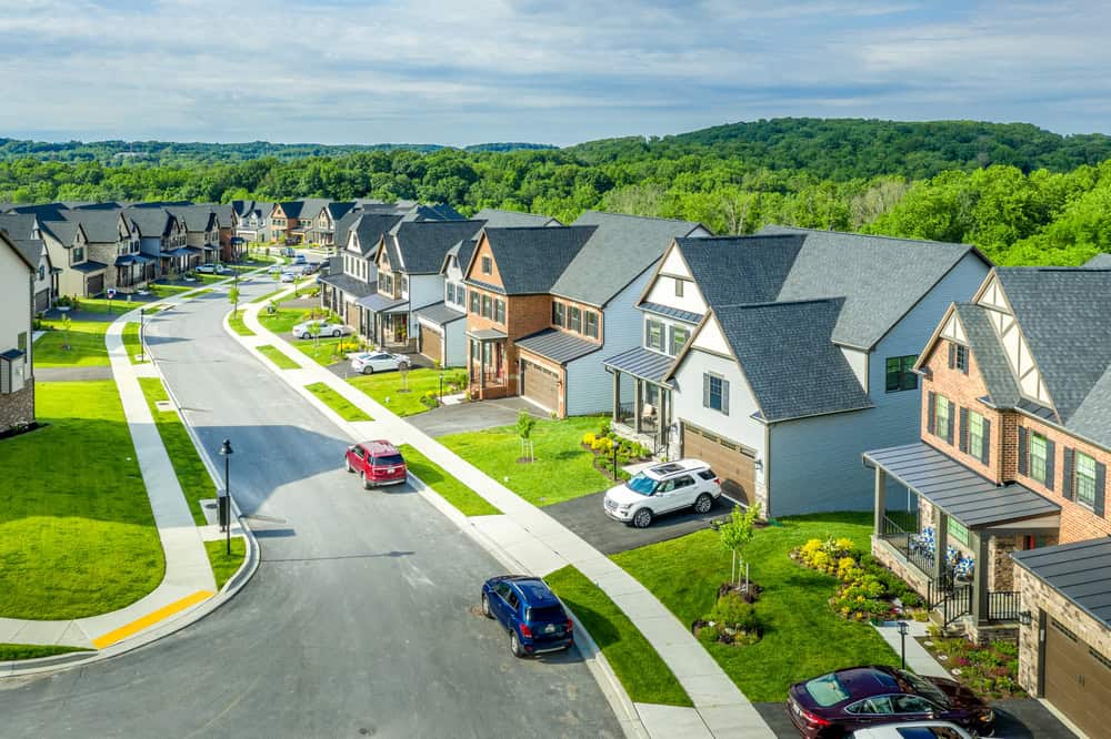 Suburban neighborhood with modern two-story homes, cars parked in driveways, and well-maintained lawns managed by diligent property managers. Aerial view reveals a winding street and lush green landscape in the background. Suburban neighborhood with modern two-story homes, cars parked in driveways, and well-maintained lawns managed by diligent property managers. Aerial view reveals a winding street and lush green landscape in the background.