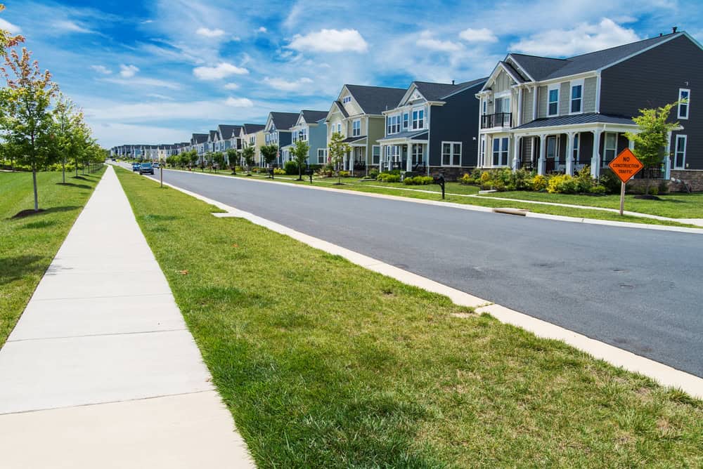 Suburban street view with a row of modern houses maintained by HOAs, a grassy lawn, and a sidewalk under a clear blue sky. A caution sign is visible on the right. Suburban street view with a row of modern houses maintained by HOAs, a grassy lawn, and a sidewalk under a clear blue sky. A caution sign is visible on the right.