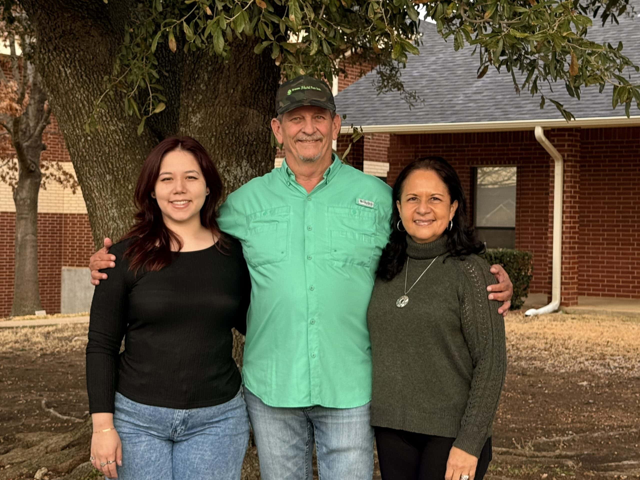 Three people stand together outside in front of a tree and a brick building, smiling at the camera—capturing the friendly spirit of Trees Hurt Too, Inc. Learn more on our About Us page.
