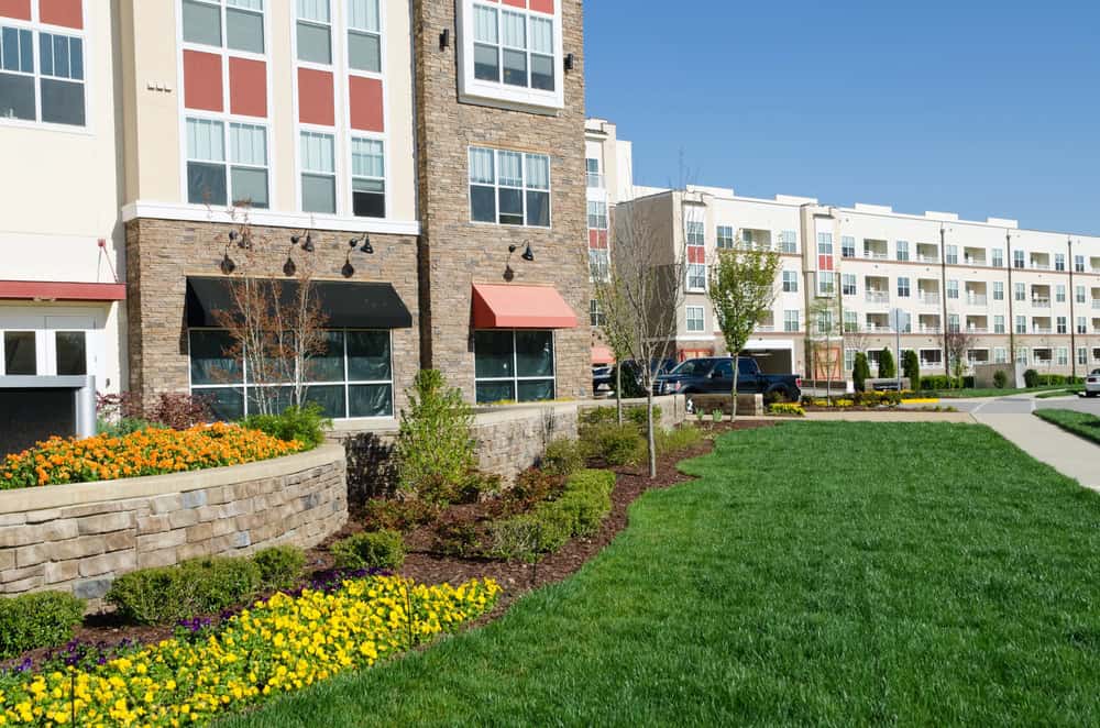 Modern apartment buildings with large windows, landscaped gardens, green lawn, and a sidewalk on a sunny day, all meticulously maintained by dedicated property managers.