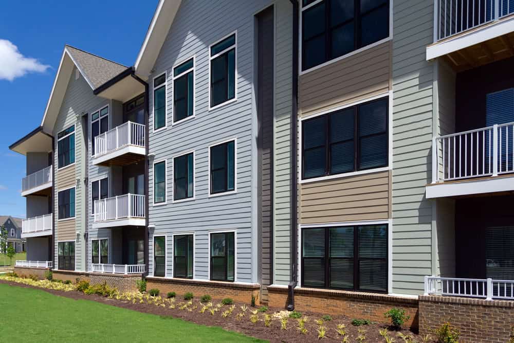Modern multi-story apartment building with light-colored siding, several balconies, large windows, and landscaped garden—perfect for property managers or HOAs seeking attractive residences under a bright blue sky.