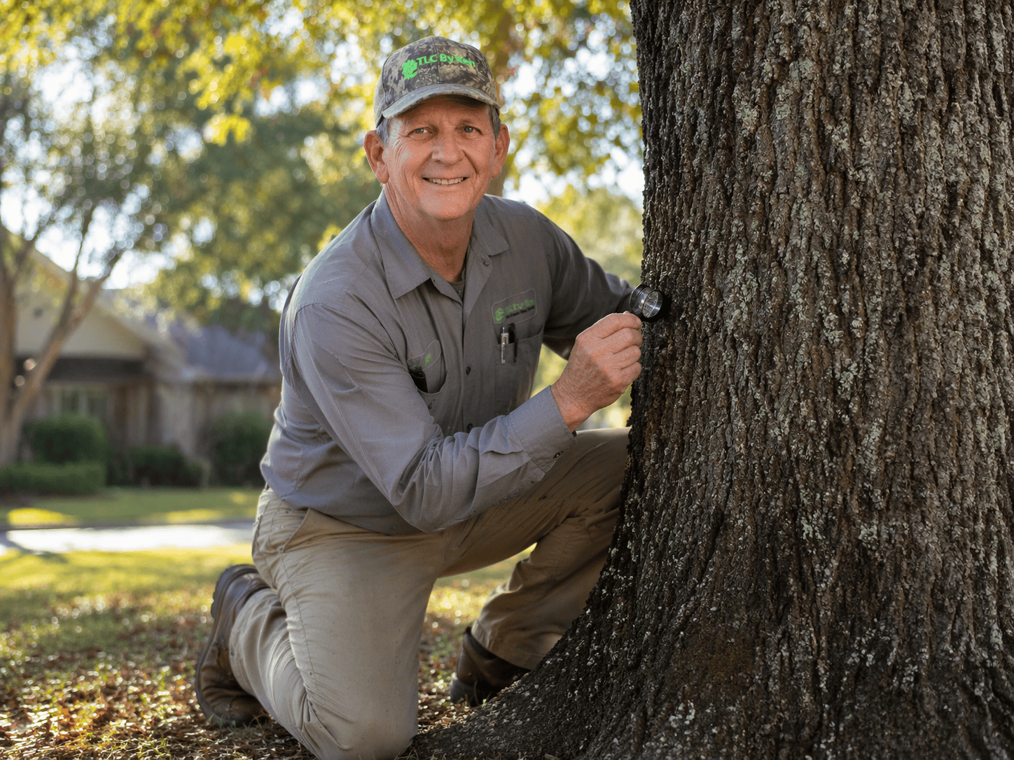 A man in work clothes and a cap kneels by a large tree, examining the trunk with a magnifying tool—your local tree doctor Fort Worth TX ensuring healthy trees in your residential area.